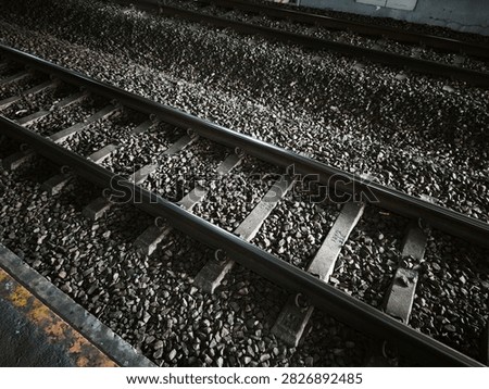 The image presents a high-angle, close-up view of multiple railway tracks running parallel to each other. Two steel rails are prominently featured in the foreground, securely fastened to light grey concrete sleepers. The entire trackbed is densely packed with dark, textured ballast stones and gravel. In the upper portion of the frame, another set of tracks is partially visible, creating a repeating pattern. The lighting highlights the top edge of the metal rails and casts deep shadows between the coarse stones, emphasizing the industrial and heavy-duty nature of the transit infrastructure.