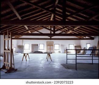 The image presents a spacious artist's studio with exposed wooden beams, white walls, and multiple canvases and art materials scattered throughout, suggesting a creative and work-intensive environment