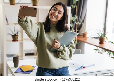Image of a pretty cute cheerful positive young girl student indoors holding clipboard take a selfie by mobile phone. - Shutterstock ID 1696378222