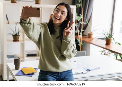 Image of a pretty cute happy positive young girl student indoors take a selfie by mobile phone showing peace gesture. - Shutterstock ID 1696378219