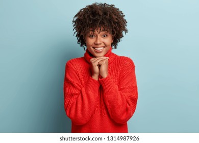 Image of pretty gentle young woman with healthy dark skin, crisp hair, keeps hands together, expresses positiveness, wears red sweater, isolated over light blue background. People and joy concept - Shutterstock ID 1314987926