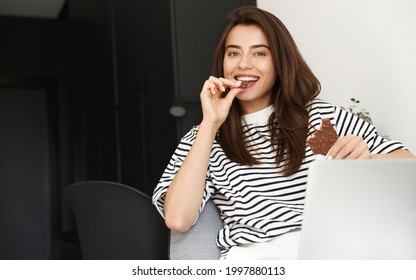 Image Of Pretty Smiling Woman Eating Chocolate Bar And Using Computer At Home. Young Woman Binge Watching Tv Series Or Movie On Laptop, Lying In Sofa With Sweets