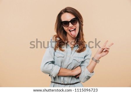 Image of pretty young happy woman wearing sunglasses standing isolated over beige background wall looking camera showing tongue and peace gesture.