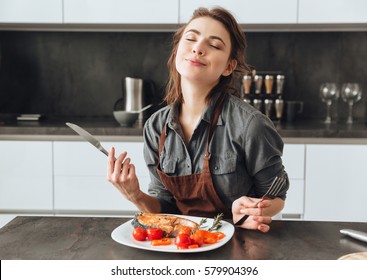 Image of pretty young woman sitting in kitchen while eating fish and tomatoes. - Shutterstock ID 579904396