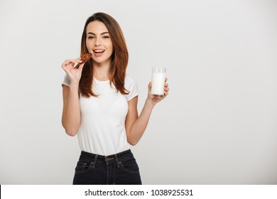 Image of pretty young woman standing isolated over grey wall background eating cookie drinking milk.