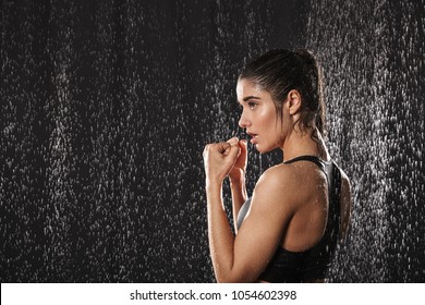 Image In Prifile Of Wet Adorable Woman Boxing And Standing In Defense Position Under Rain Drops Isolated Over Black Background