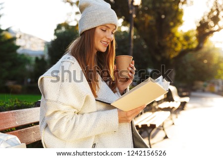 Image of a redhead woman sitting on bench outdoors holding reading book drinking coffee.
