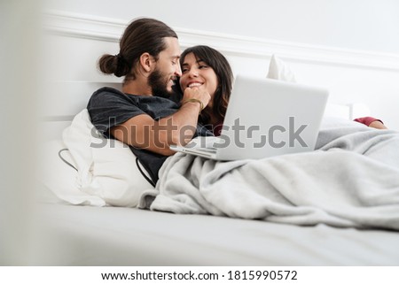 Image of romantic cheerful couple using laptop and hugging while lying in bed at home