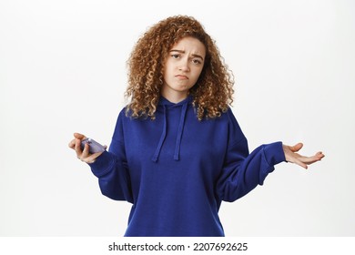 Image Of Sad And Confused Young Woman Shrugging With Mobile Phone, Looking Upset And Uneasy, Standing Over White Background In Blue Hoodie.