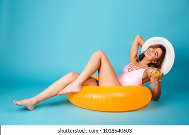 Image of a satisfied girl dressed in swimsuit sitting on inflatable ring with a cocktail and relaxing isolated over blue background