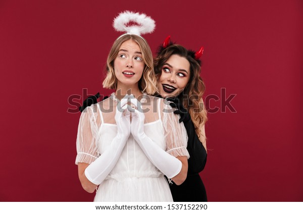 Image of scary woman\
demon near thinking angel in carnival costumes isolated over red\
wall background.