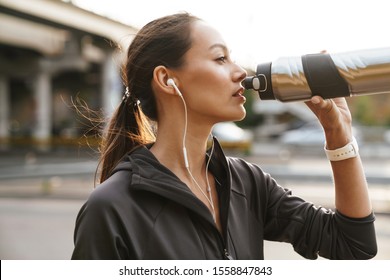 Image of serious athletic woman using earphones and drinking water while working out near road bridge in morning - Powered by Shutterstock