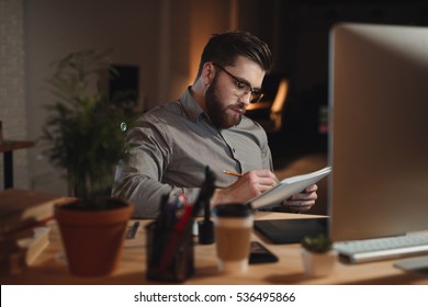 Image Of Serious Bearded Web Designer Dressed In Shirt Working Late At Night And Looking At Notebook While Writing Notes.