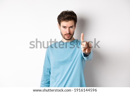 Image of serious young man with beard, shaking finger in disapproval, prohibit or forbid something, standing over white background and saying no
