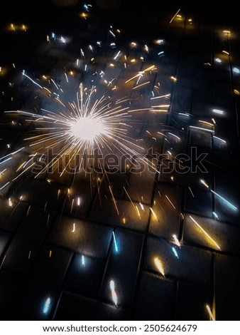 The image showcases a ground-level view of a firework, likely a ground spinner or fountain, in the midst of its display. Bright, colorful sparks burst outward from the center, illuminating the dark.