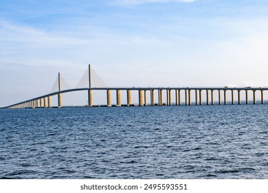 Image showcasing a long, modern cable-stayed bridge spanning over a vast body of water. The bridge features tall pylons and multiple support columns, stretching across the horizon.