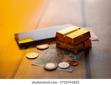 Image showing stack of gold bars sits on a GHS 100 banknote and various coins, next to a black smartphone, all resting on a wooden surface, depicting themes of wealth and digital finance.