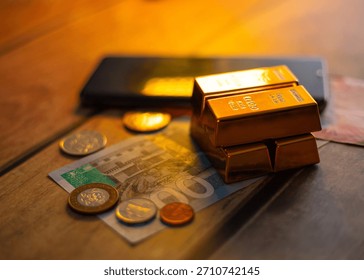 Image showing stack of gold bars sits on a GHS 100 banknote and various coins, next to a black smartphone, all resting on a wooden surface, depicting themes of wealth and digital finance.