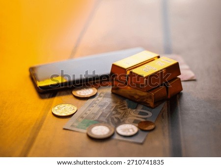 Image showing stack of gold bars sits on a GHS 100 banknote and various coins, next to a black smartphone, all resting on a wooden surface, depicting themes of wealth and digital finance.