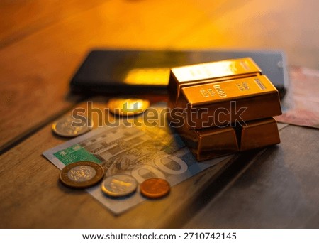 Image showing stack of gold bars sits on a GHS 100 banknote and various coins, next to a black smartphone, all resting on a wooden surface, depicting themes of wealth and digital finance.