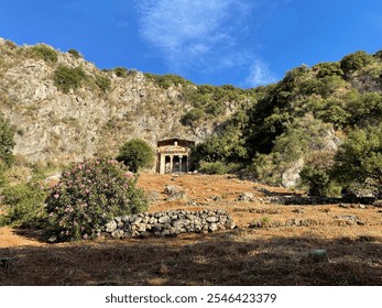 The image shows the ancient city of Telmessos, featuring a rock-carved Lycian tomb surrounded by rugged cliffs, green foliage, and pink flowering bushes under a bright blue sky.  - Powered by Shutterstock