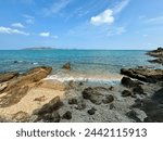 The image shows a beach with rocks . The sky is bright blue, and there are fluffy white clouds in the distance. There are waves crashing on the shore, and there are rocks scattered along the beach