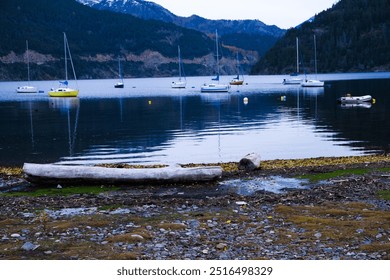 The image shows a calm lake with sailboats anchored near the shore. The water reflects the boats and surrounding mountains. The foreground features rocky terrain with logs, creating a serene lakeside  - Powered by Shutterstock