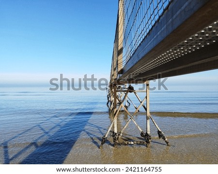 The image shows a calm and relaxing scene of a clear blue sea with a long pier stretching out into the water. The sky is a cloudless blue, and the sun is shining brightly. Photo take in Rimini