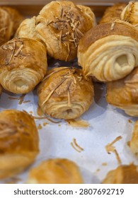 The image shows a close-up, top-down view of several golden-brown pastries arranged on white parchment paper. These appear to be cheese croissants or cheese rolls (often referred to as croissant keju or bolen style pastries).
​Key Details:
​Appearance and Texture: The pastries are rolled up, showcasing a flaky, laminated puff pastry texture with visible layers on the open sides. The tops have a glossy, golden-brown baked finish.
​Toppings and Filling: They are garnished with scattered shreds of toasted, crispy cheese on top. A pale, creamy filling (likely cheese or custard) is slightly visible from the rolled openings on the sides.
​Composition: The pastries are placed together in a bakery display or tray. The photo uses a shallow depth of field, keeping the main croissants in the center and upper right sharp, while those in the foreground and edges are softly out of focus.
