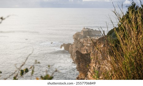 The image shows a coastal cliff with a fortress perched at the edge, overlooking the ocean. Waves crash against the rocky shore, framed by vegetation in the foreground, creating a dramatic contrast. - Powered by Shutterstock