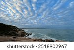 The image shows a coastal path along rocky shores, with the ocean stretching to the horizon. Fluffy clouds fill the blue sky, while distant islands peek through. A serene, natural coastal scene.