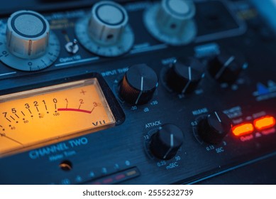 The image shows a detailed view of an audio mixing console, featuring various knobs, dials, and a glowing VU meter, indicating audio levels in a studio setting. - Powered by Shutterstock