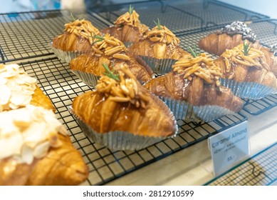 The image shows a display of assorted baked pastries including croissants with toppings, arranged on a wire rack in a bakery or pastry shop setting, highlighting fresh, flaky, golden-brown treats suitable for breakfast or snack time.