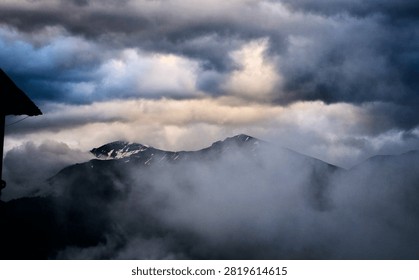 The image shows a dramatic, moody mountain landscape with rugged, snow-dusted twin peaks partially shrouded in dense, low-hanging layers of thick white fog and heavy, dark, overcast storm clouds above