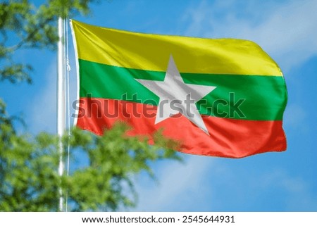 The image shows the flag of Myanmar, featuring yellow, green, and red stripes with a white star. The Myanmar flag waves against a clear blue sky. National flag of Myanmar.