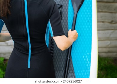 The image shows a girl in a wetsuit holding a blue surfboard, ready for surfing. Greenery and wooden wall in the background - Powered by Shutterstock