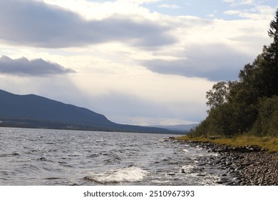 The image shows a lakeside view with choppy water, a rocky shore, and mountains in the distance under a cloudy sky, creating a sense of tranquility and natural beauty. - Powered by Shutterstock