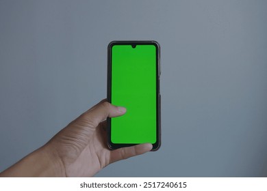 The image shows a left hand holding a smartphone with a bright green screen, a plain gray background. The green screen can be used to display changeable content. - Powered by Shutterstock