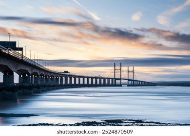 The image shows a long, elegant suspension bridge over calm waters during sunset or sunrise. The sky is filled with soft, streaky clouds, and the fading light casts a serene, blueish hue over  - Powered by Shutterstock