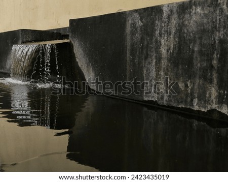The image shows a  man-made mini waterfall with a black painted wall, capturing the man made beauty of the scene. The water cascades down the wall, creating a beautiful reflection in the pool below.