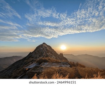 The image shows a mountain peak at sunset. The sky is a mix of blue and light orange, with some clouds. The sun is setting behind the mountain, casting a warm glow over the scene. - Powered by Shutterstock