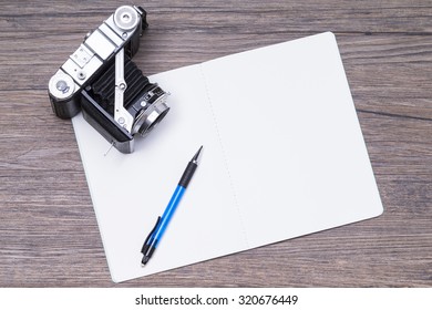 Image shows a notepad with pen and camera on a wooden table