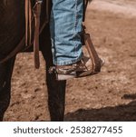 The image shows a person in blue jeans and tan boots with silver spurs on horseback, one foot in a brown leather stirrup. The ground is dusty, and the scene has a rustic, Western vibe.