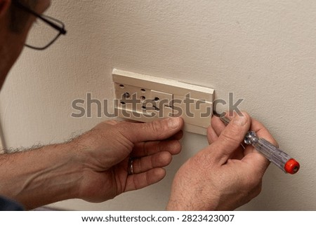 The image shows a person engaged in the process of repairing an electrical outlet using a polarity tester. The individual is working with a toolkit, specifically focusing on one piece that appears to be in the process of being attached or disassembled from the outlet. The context of the image suggests it may be taken in a domestic setting and captures a moment of maintenance or DIY electrical work.