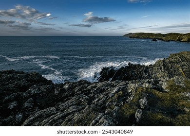 The image shows a rocky shore with the ocean stretching out before it. The sky is a bright blue, with some clouds visible. The water is a deep blue color, and the waves are breaking on the shore. - Powered by Shutterstock