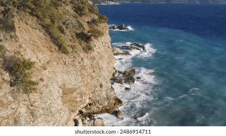 The image shows a rugged coastal cliff with trees and shrubs growing on it, overlooking a deep blue sea. The waves crash against the rocky shoreline, creating white foam. The clear, turquoise waters n - Powered by Shutterstock