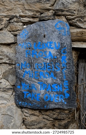 The image shows a rustic stone wall with a mounted plaque bearing a multilingual message about environmental responsibility. The blue-painted text, written in Spanish, Catalan, and English, calls for visitors not to litter and to take their trash with them. The scene evokes civic awareness, respect for nature, and the quiet dignity of communal care in a mountainous region.