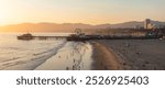 The image shows Santa Monica Pier at sunset, featuring the Ferris wheel, beachgoers, and the Santa Monica Mountains with the cityscape in the background.