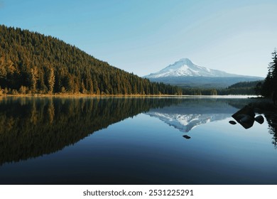 The image shows a serene landscape with a calm lake reflecting a forested hillside and a snow-capped mountain. The clear sky enhances the sense of tranquility and pristine beauty of the scene. - Powered by Shutterstock