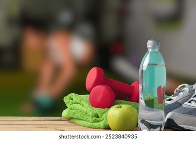 The image shows a set of gym essentials arranged on a table, including a pair of small red dumbbells, a green towel, a bottle of water, a green apple, and a pair of gray sneakers. - Powered by Shutterstock